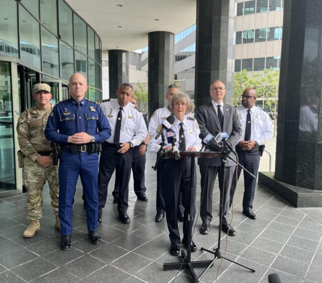 Drop in crime Group of uniformed officials and a speaker at a press conference outside a modern building with microphones on a stand in front of them