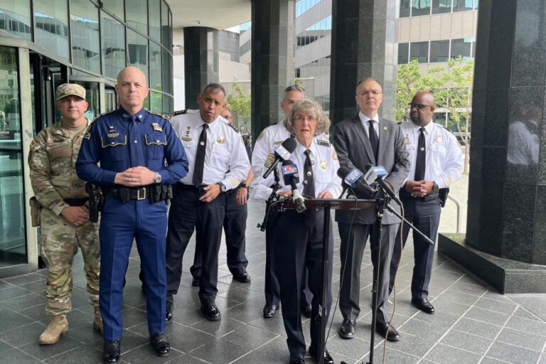 Drop in crime Group of uniformed officials and a speaker at a press conference outside a modern building with microphones on a stand in front of them