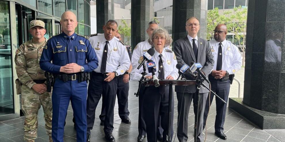 Drop in crime Group of uniformed officials and a speaker at a press conference outside a modern building with microphones on a stand in front of them